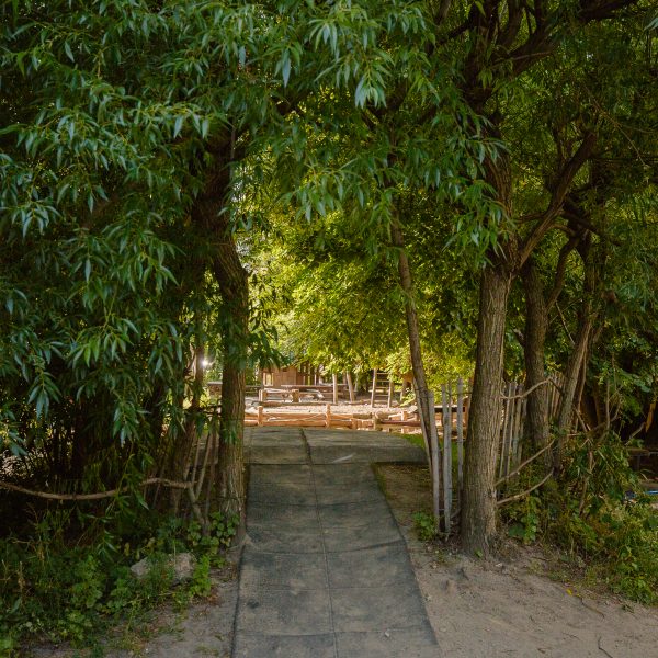 Kita-Garten mit Sand und Gehplatten, umringt von grünen Bäumen.