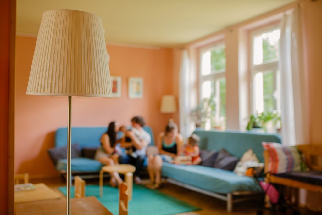 „Wer betreut mein/unser Kind?“ Die Wahl zwischen Kindertagespflege oder Kinderkrippe – Info- & Fragerunde - FiZ Leipzig Mütter und Väter sitzen mit ihren Kindern auf zwei Sofas im offenen Treff des Familienzentrums, die abgebildeten Personen sind unscharf zu erkennen