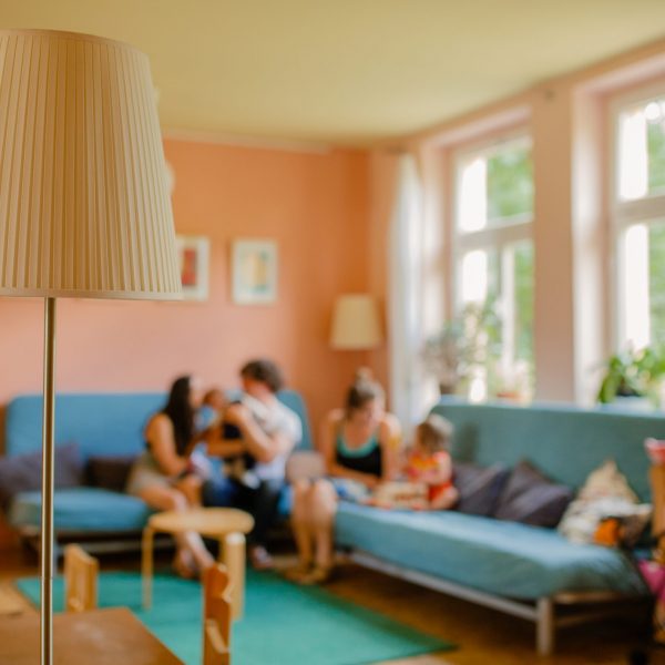 Mütter und Väter sitzen mit ihren Kindern auf zwei Sofas im offenen Treff des Familienzentrums, die abgebildeten Personen sind unscharf zu erkennen