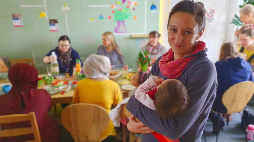 Im Vordergrund hält eine Frau ein Baby im Arm und lächelt uns zu, Im Hintergrund sitzen Frauen am Tisch und frühstücken