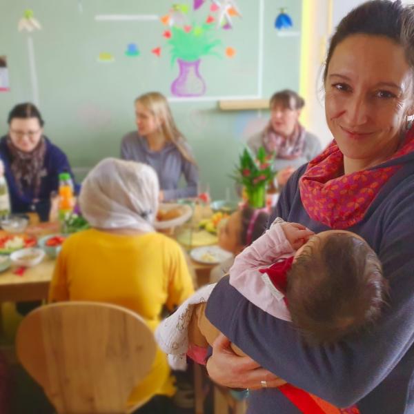 Im Vordergrund hält eine Frau ein Baby im Arm und lächelt uns zu, Im Hintergrund sitzen Frauen am Tisch und frühstücken