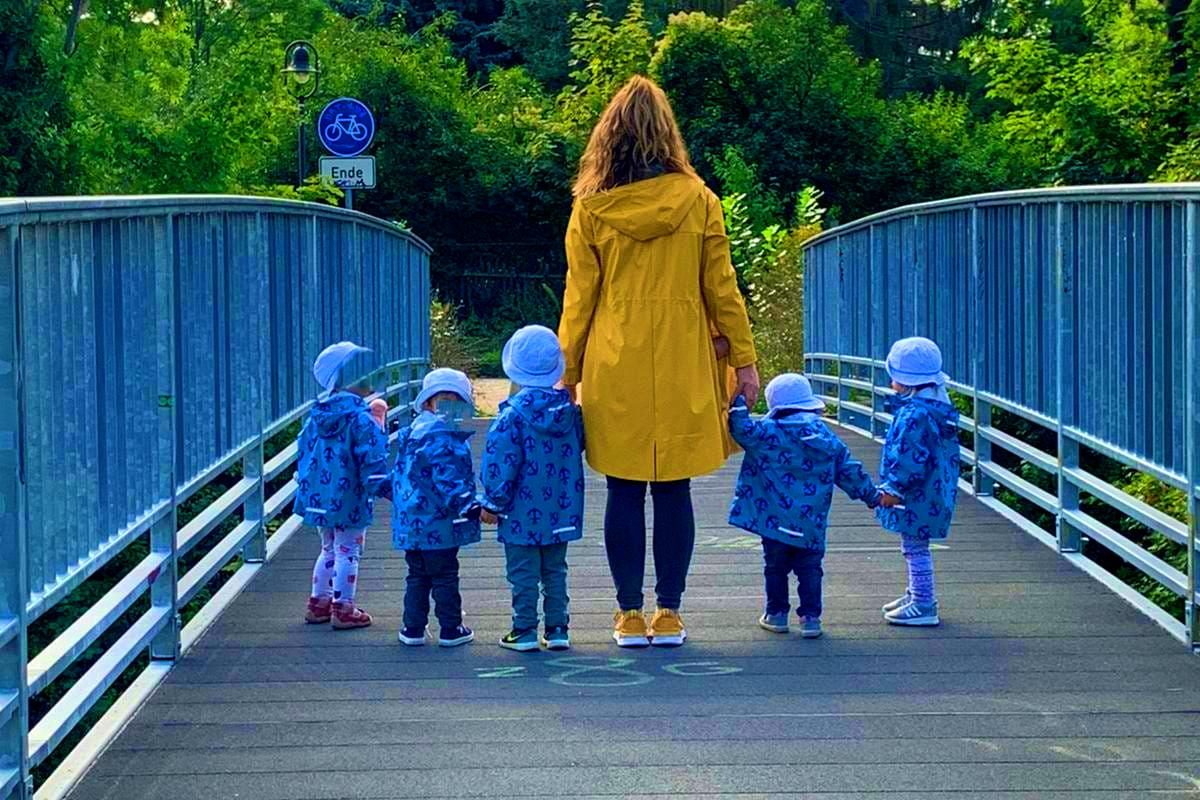 Eine Frau in gelber Jacke steht mit fünf Kindern, jeweils in blau gekleidet, auf einer Brücke.