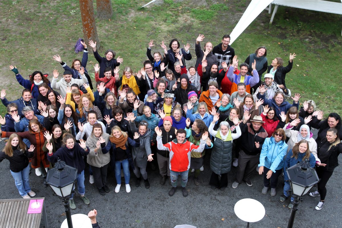 Ein Gruppenfoto der Mitarbeitenden des Mütterzentrum e.V. Leipzig, aufgenommen aus der Vogelperspektive: Personen unterschiedlichen Alters Lachen und Winken in die Kamera.
