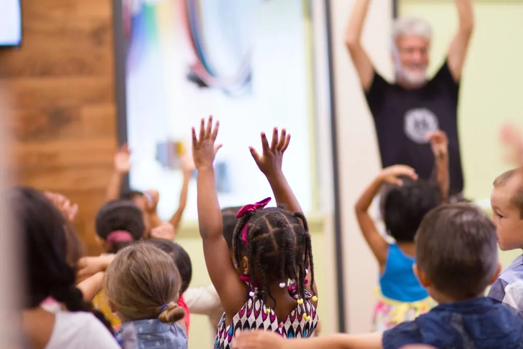 Ein Mann im Hintergrund und Kinder im Vordergrund strecken die Arme in die Höhe.