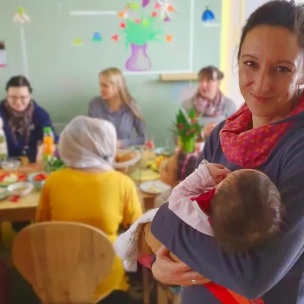 Im Vordergrund hält eine Frau ein Baby im Arm und lächelt uns zu, Im Hintergrund sitzen Frauen am Tisch und frühstücken