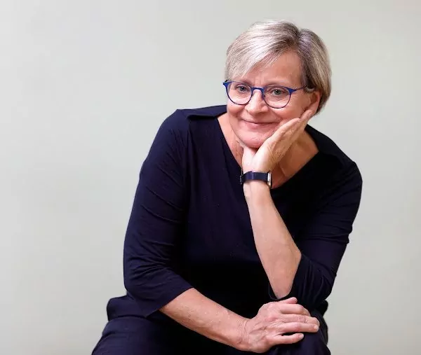 Eine Portraitaufnahme der Hebamme Raja Rohrwacher, die freundlich in die Kamera lächelt. Sie trägt kurzes graues Haar, ein dunkles Kleid und eine Brille mit dunklem Rahmen.