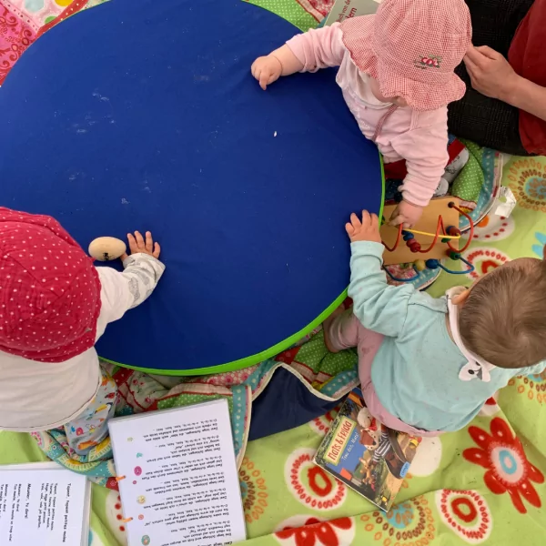 Das Bild zeigt eine Impression von oben auf eine Spielsituation für Kleinkinder. Es sind deri Kleinkinder, Spielzeuge, Liedtexte zu sehen.
